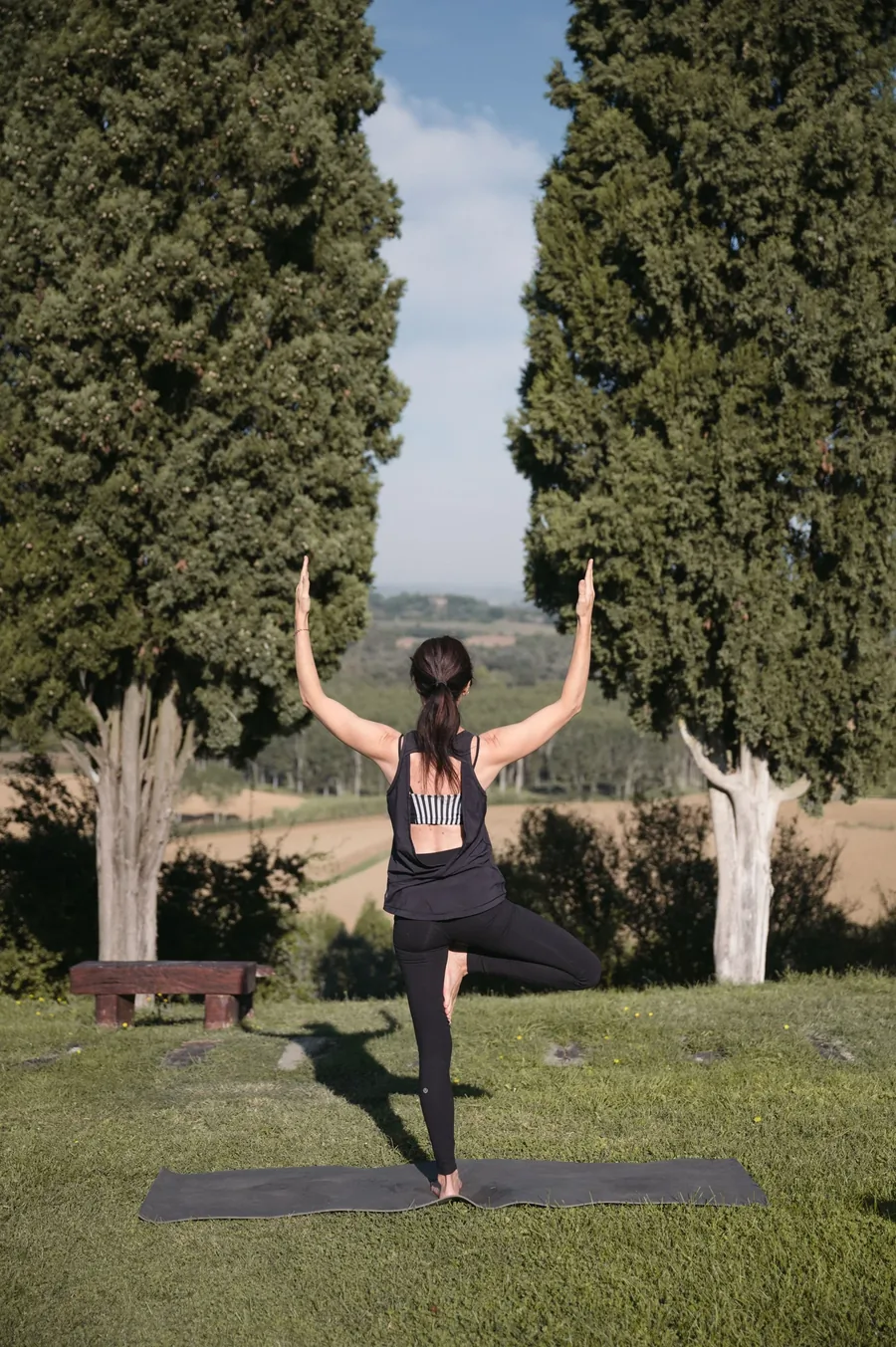 A woman in tree pose between two cypresses, overlooking the valley