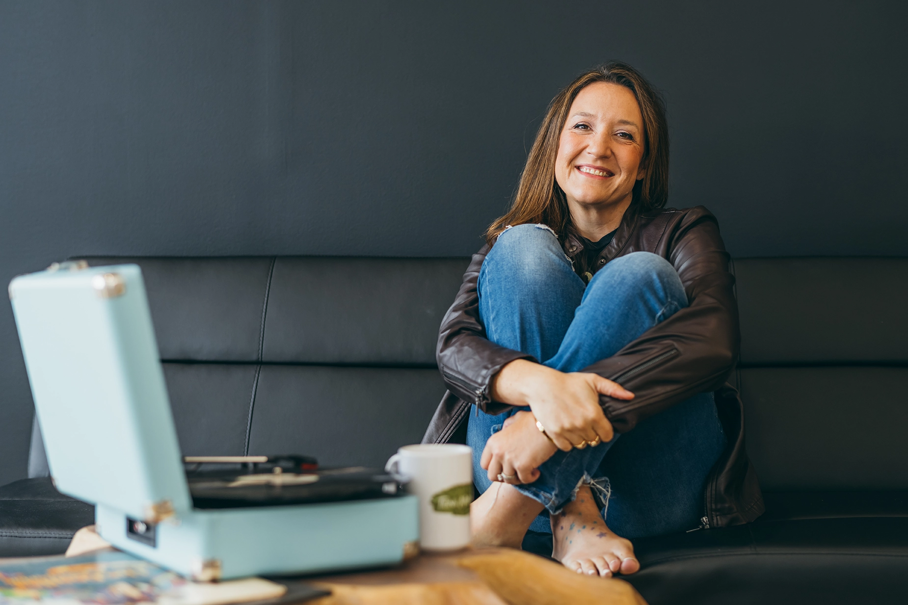 A woman in a brown leather jacket and jeans sits barefoot on a black couch, smiling with her arms around her knees. A blue record player and a coffee mug are on the wooden table in front of her.